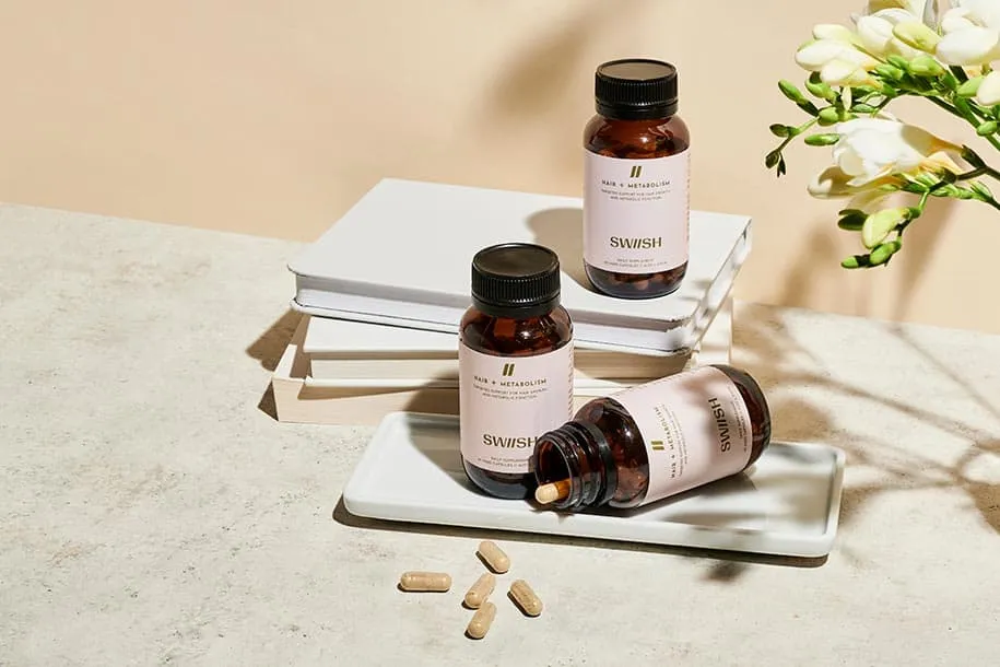Three bottles of SWIISH Hair + Metabolism supplements styled with a stack of books and fresh flowers