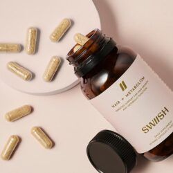 Bottle of SWIISH Hair + Metabolism supplement with lid open and capsules spilling out on a beige background